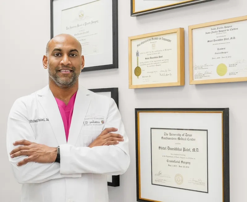 El Dr. Shitel D. Patel con bata blanca y los brazos cruzados, frente a sus diplomas enmarcados